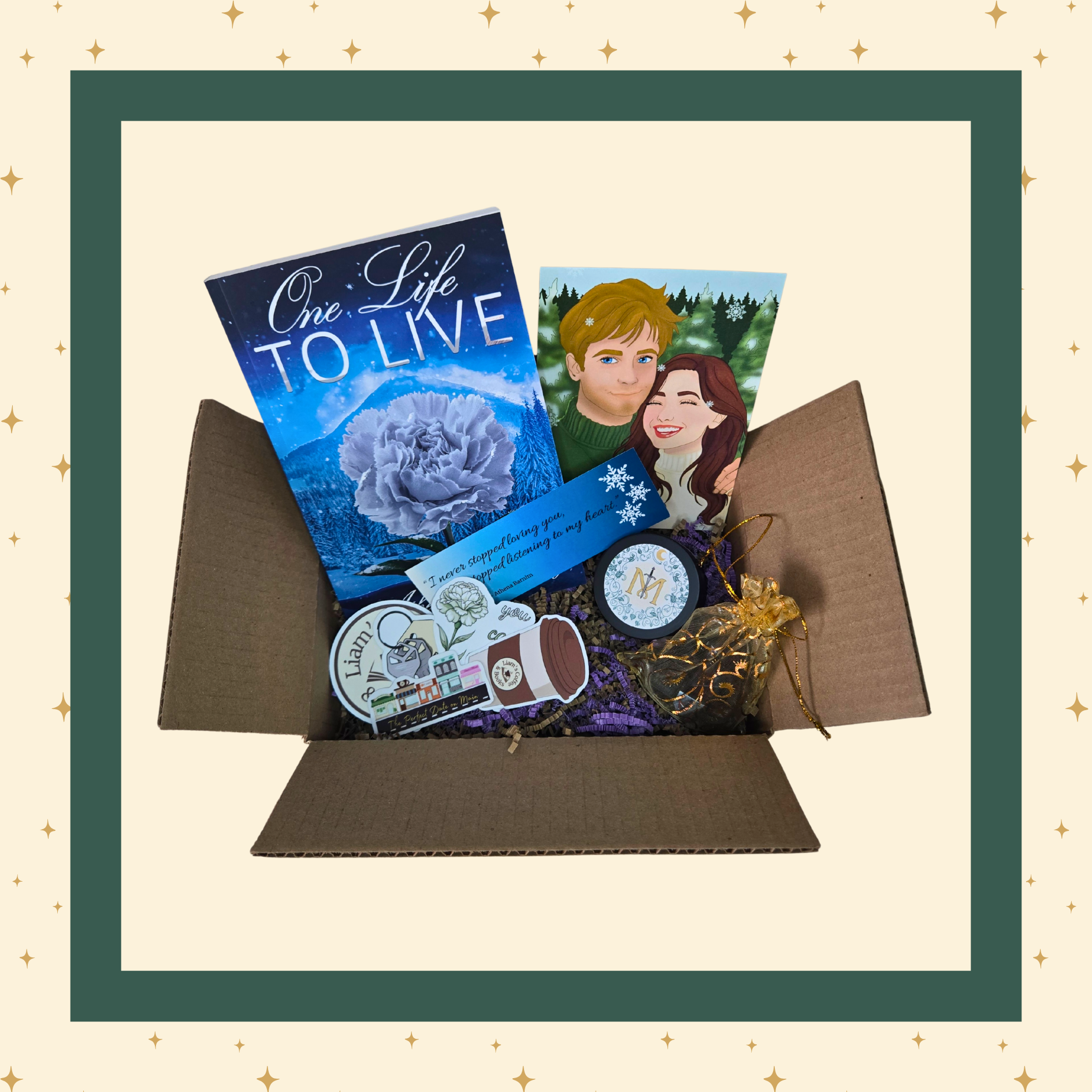 An open gift box containing a curated selection of items: a blue-covered book titled "One Life To Live," art featuring the main characters of the book, a round candle container with a stylized "M" label, a set of resin polyhedral dice, several themed stickers, and additional small items.