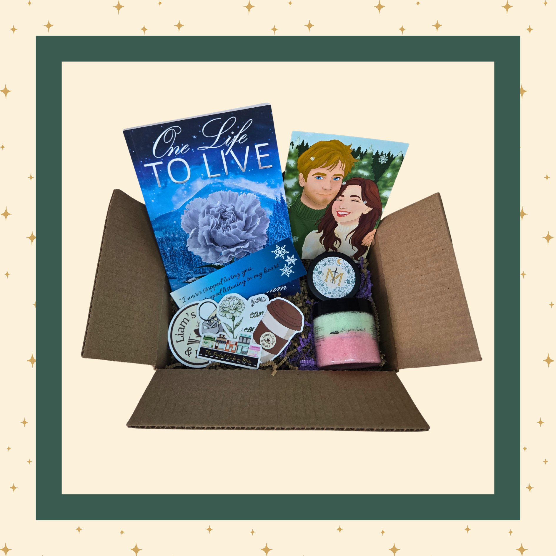 An open gift box containing a curated selection of items: a blue-covered book titled "One Life To Live," art featuring the main characters of the book, a round candle container with a stylized "M" label, a multicoloured sugar scrub, several themed stickers, and additional small items.
