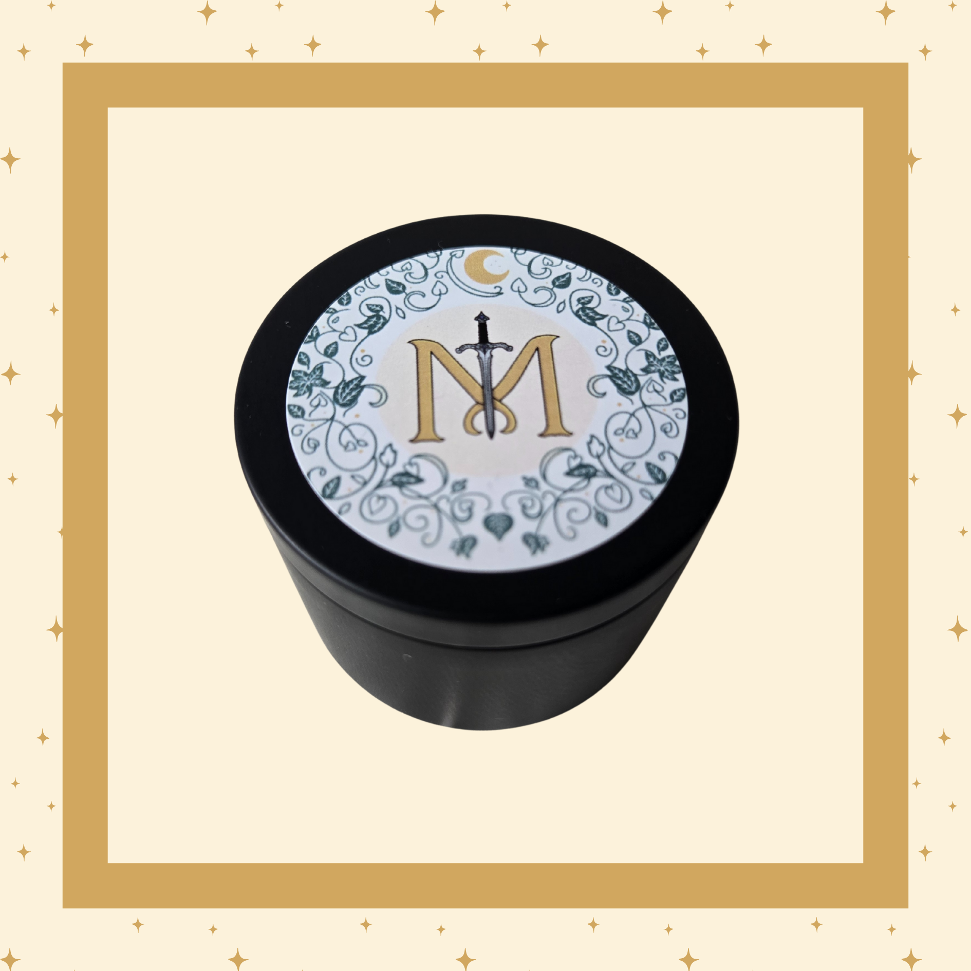 A small black candle container with a circular decorative lid. The lid features a gold letter "M" intersected by a silver sword, surrounded by green vines and leaves, with a gold crescent moon at the top.