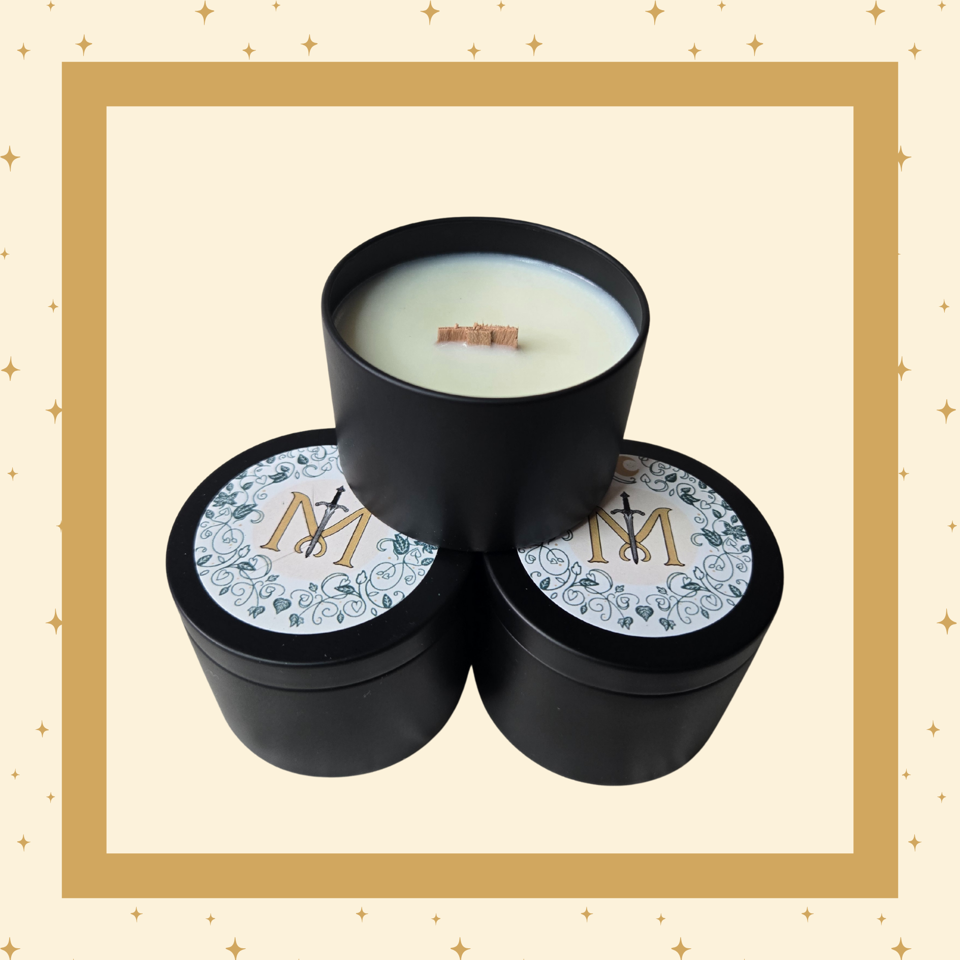 Three black candle containers arranged in a pyramid. Two are closed with decorative lids featuring a gold "M" intersected by a black sword, surrounded by green vines. The third container is open, showing white wax and a centred wooden wick.